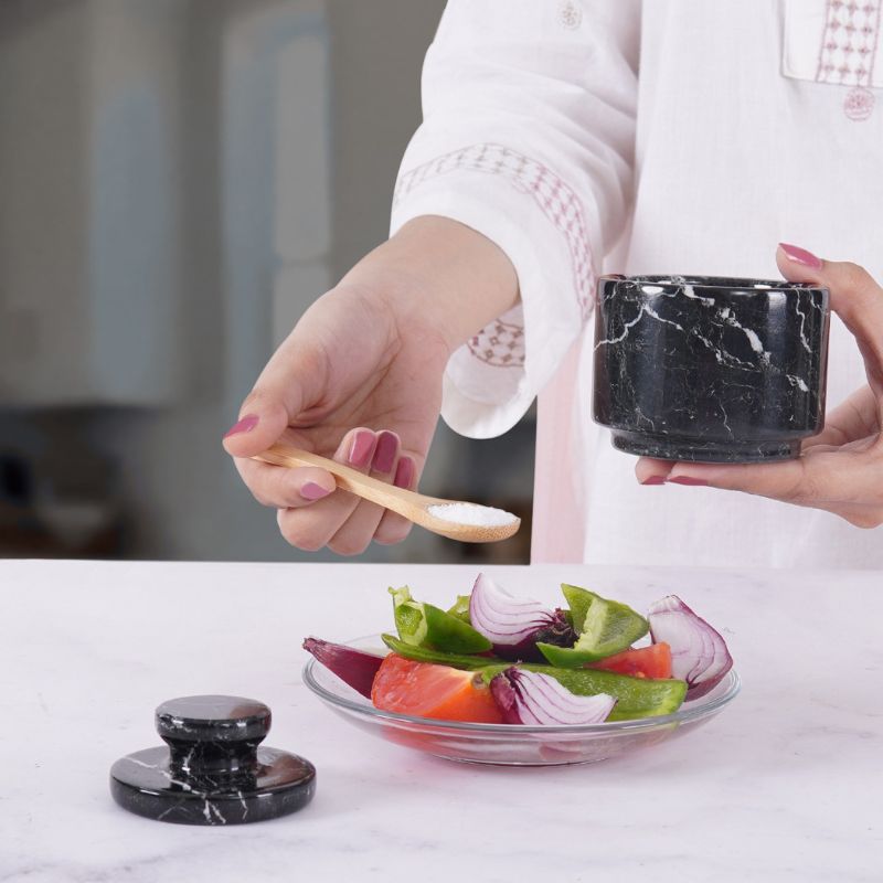 Woman using black marble salt cellar with spoon to season fresh salad